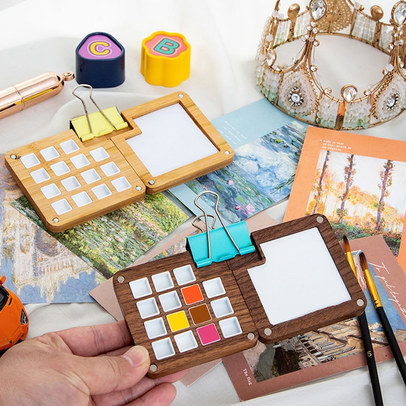 Op de afbeelding: Twee houten aquarelpaletten met verwisselbare bakjes. De paletten zijn open om de lege bakjes en een blanco wit papieroppervlak te laten zien. De paletten zijn gemaakt van licht en donker hout. Een palet heeft een blauwe clip en de andere een gele clip.