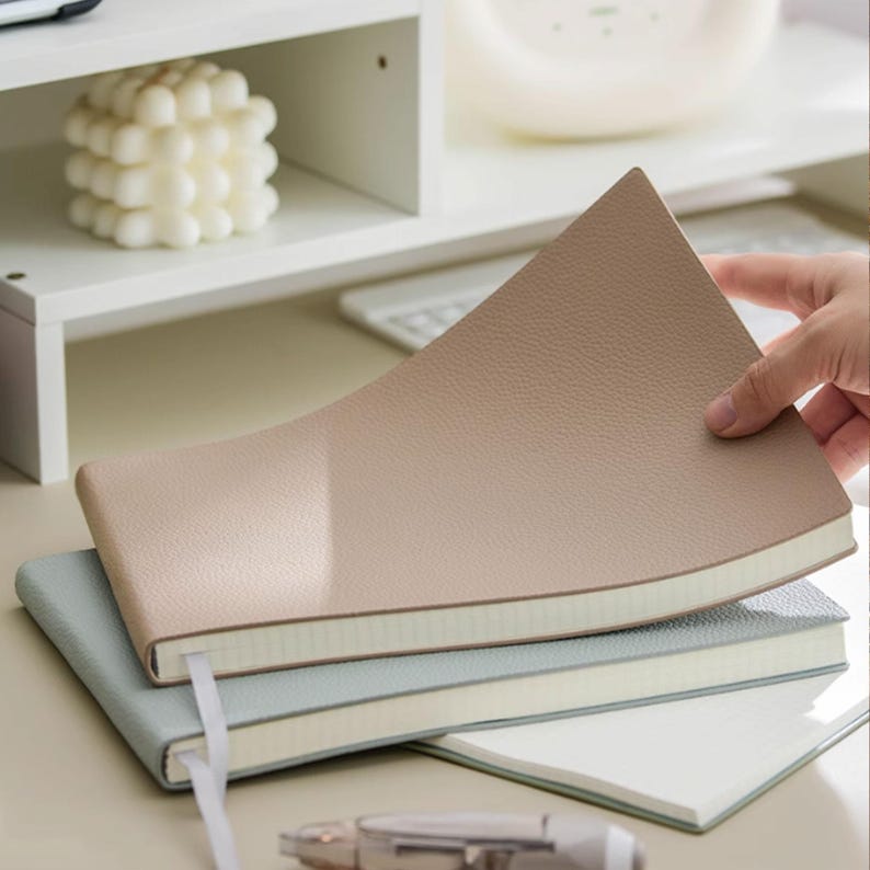 May include: A stack of three notebooks in neutral tones. The top notebook is a light tan color, with a textured cover. The middle notebook is a light blue, and the bottom notebook is white. Each notebook has a ribbon bookmark.