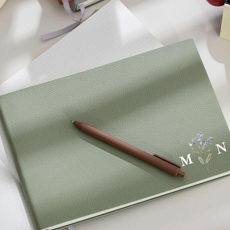 May include: A sage green journal with a textured cover, a brown pen, and a white grid paper. The journal is decorated with a floral design and the letters "M" and "N". The pen is resting on the journal.