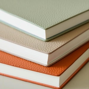 May include: A stack of three notebooks with textured covers in sage green, tan, and orange. The notebooks have white paper and a clean, minimalist design, perfect for journaling or note-taking.