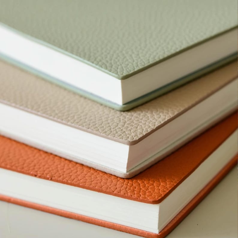 May include: A stack of three notebooks with textured covers in sage green, tan, and orange. The notebooks have white paper and a clean, minimalist design, perfect for journaling or note-taking.