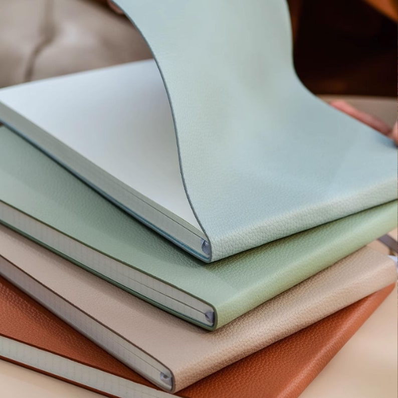 May include: A stack of notebooks in various colors: light blue, green, beige, and brown. The top notebook's cover is lifted, revealing white paper. The covers have a textured, leather-like appearance, suggesting a high-quality product.