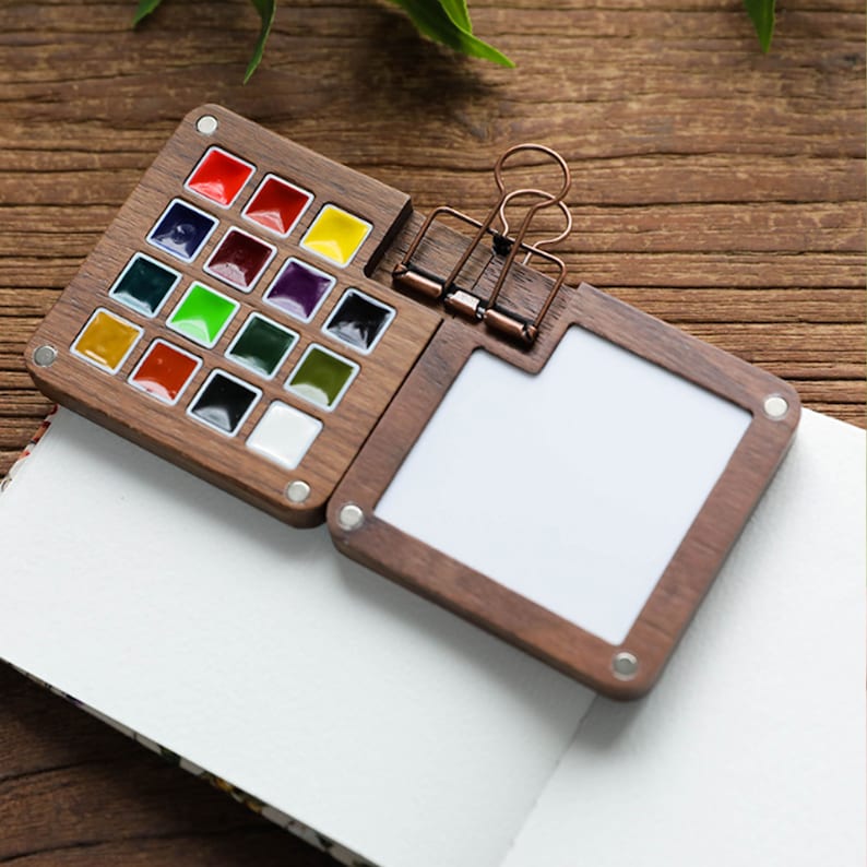 Op de afbeelding: Een houten aquarelverfset met een magnetische sluiting. De set bevat 16 aquarelverfpannetjes in verschillende kleuren, waaronder rood, oranje, geel, groen, blauw, paars en bruin. De set bevat ook een klein, blanco, wit papierblok.