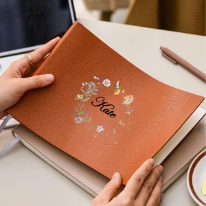 May include: A brown leather journal with the name "Kate" in script, surrounded by a floral wreath. The journal is open, revealing blank pages. The cover has a textured appearance.