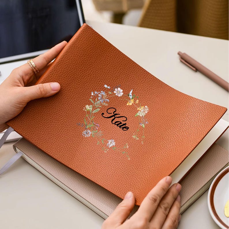 May include: A brown leather journal with the name "Kate" in script, surrounded by a floral wreath. The journal is open, revealing blank pages. The cover has a textured appearance.