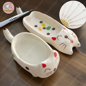 May include: Two white ceramic cat-shaped paint palettes. One palette has a colourful paint splatter design and the other is plain. Both palettes have a small, round depression for holding paint.