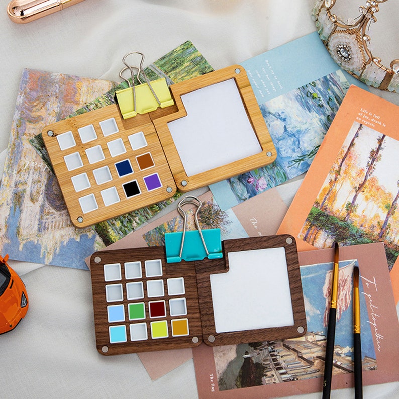 Op de afbeelding: Twee houten aquarelpaletten met een scharnierend deksel en een uitneembare lade voor verfpotjes. De palette aan de linkerkant is gemaakt van licht hout en heeft 16 verfpotjes. De palette aan de rechterkant is gemaakt van donker hout en heeft 16 verfpotjes. Beide paletten hebben een wit papierblok om verf te mengen.