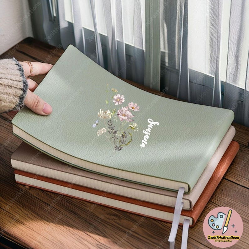 May include: A stack of notebooks with a sage green cover featuring a floral design and the word "Sugena" in white script. The notebooks are stacked on a wooden surface, with a hand holding the top notebook. The other notebooks have brown and beige covers.