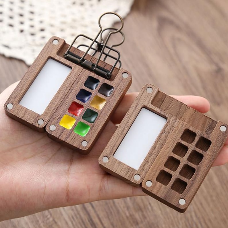 Op de afbeelding: Een houten aquarelpalette met een scharnierend deksel en een metalen clip. Het palet heeft 12 vakjes voor verf en een klein gebied om te mengen. Het palet is open en toont een verscheidenheid aan kleuren aquarelverf.