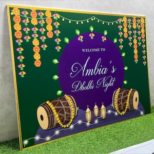 May include: A green and purple welcome sign with the text "Welcome to Ambia's Dholki Night". The sign is decorated with yellow and orange flowers, green and pink tassels, and string lights. There are two brown drums with yellow beads on them and three gold lanterns.