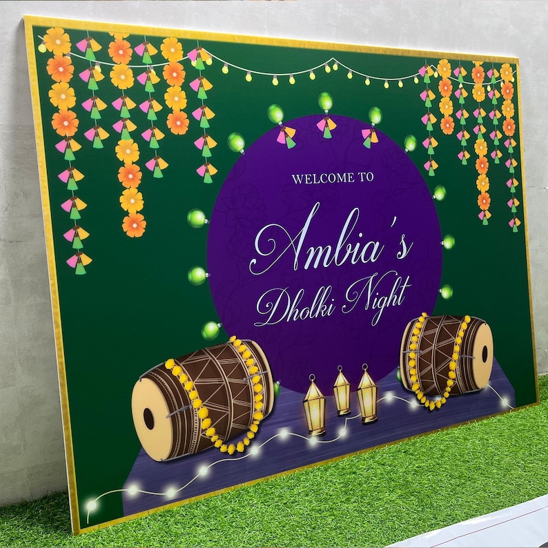 May include: A green and purple welcome sign with the text "Welcome to Ambia's Dholki Night". The sign is decorated with yellow and orange flowers, green and pink tassels, and string lights. There are two brown drums with yellow beads on them and three gold lanterns.