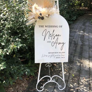 Puede incluir: Un cartel de boda blanco con texto negro anunciando la boda de Nelson y Amy. El cartel tambi&eacute;n incluye la fecha 20.05.2023 y la ubicaci&oacute;n Riverview Island. El cartel est&aacute; en un caballete blanco.