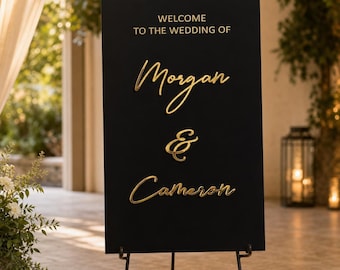 Puede incluir: Un letrero de bienvenida de boda rectangular negro con letras doradas en 3D que dicen "WELCOME TO THE WEDDING OF Morgan & Cameron". El letrero está montado sobre un caballete de metal negro con adornos de volutas en la parte superior e inferior.