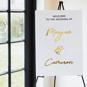 Puede incluir: Se&ntilde;al de bienvenida de boda blanca con letras doradas. La se&ntilde;al dice "Bienvenido a la boda de Morgan & Cameron".