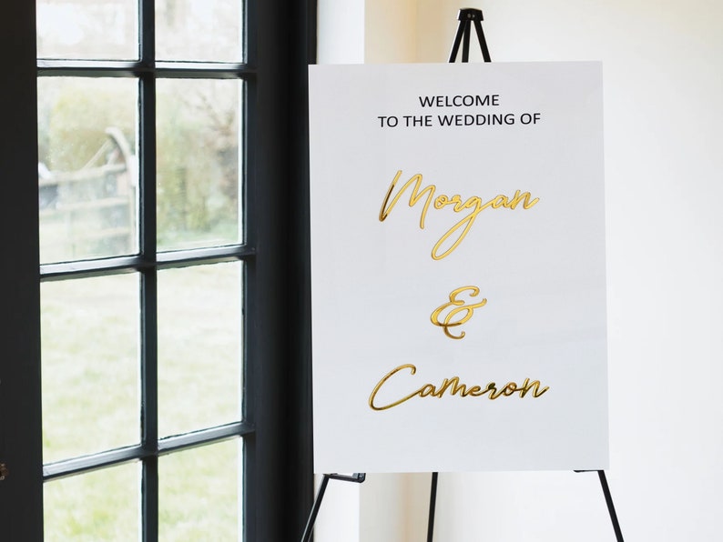Puede incluir: Se&ntilde;al de bienvenida de boda blanca con letras doradas. La se&ntilde;al dice "Bienvenido a la boda de Morgan & Cameron".