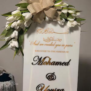 Puede incluir: Un cartel de boda blanco con caligraf&iacute;a dorada. El cartel dice "Y te creamos en parejas." y "Bienvenido al Nikkah de Mohamed & Yousra".