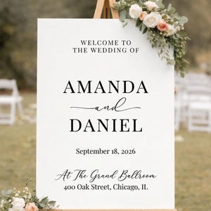 May include: A white wedding welcome sign on a wooden easel, adorned with floral arrangements. The sign reads "WELCOME TO THE WEDDING OF AMANDA and DANIEL" with the date "September 18, 2026" and venue details.