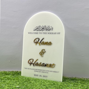 Puede incluir: Un cartel blanco con la parte superior arqueada, que presenta la frase &aacute;rabe "Bismillah ar-Rahman ar-Rahim" en caligraf&iacute;a negra. El texto "Welcome to the Nikkah of Hana & Hasanat" est&aacute; escrito en negro debajo de la caligraf&iacute;a. La cita "And he placed between you love and mercy" est&aacute; escrita en negro debajo de los nombres, seguida de "Surah Ar-Rum 30:21" en negro. La fecha "May 10, 2025" est&aacute; escrita en negro en la parte inferior del cartel.