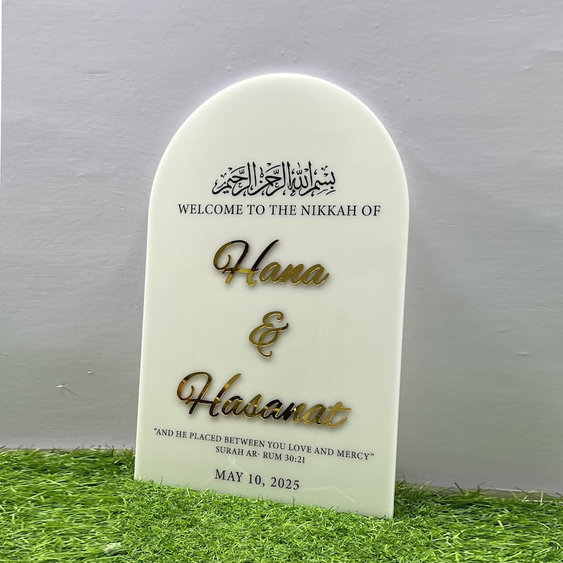 Puede incluir: Un cartel blanco con la parte superior arqueada, que presenta la frase &aacute;rabe "Bismillah ar-Rahman ar-Rahim" en caligraf&iacute;a negra. El texto "Welcome to the Nikkah of Hana & Hasanat" est&aacute; escrito en negro debajo de la caligraf&iacute;a. La cita "And he placed between you love and mercy" est&aacute; escrita en negro debajo de los nombres, seguida de "Surah Ar-Rum 30:21" en negro. La fecha "May 10, 2025" est&aacute; escrita en negro en la parte inferior del cartel.