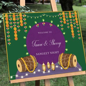May include: A welcome sign for a Sangeet Night, featuring a purple circle with the text "Welcome to Tanvi & Sherry Sangeet Night". The background is green with garlands of flowers and string lights. There are two brown drums with yellow beads on the bottom of the sign.