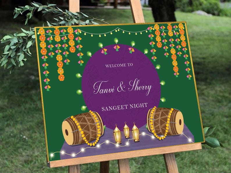 May include: A welcome sign for a Sangeet Night, featuring a purple circle with the text "Welcome to Tanvi & Sherry Sangeet Night". The background is green with garlands of flowers and string lights. There are two brown drums with yellow beads on the bottom of the sign.