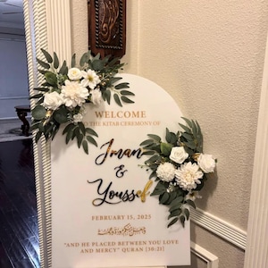 Puede incluir: Un cartel de bienvenida blanco con letras doradas para una ceremonia de boda. El cartel dice "Bienvenido a la ceremonia del Kitab de Iman & Youssef, 15 de febrero de 2025." El cartel tambi&eacute;n incluye caligraf&iacute;a &aacute;rabe y una cita del Cor&aacute;n: "Y &Eacute;l puso entre vosotros el amor y la misericordia." Cor&aacute;n [30:21].