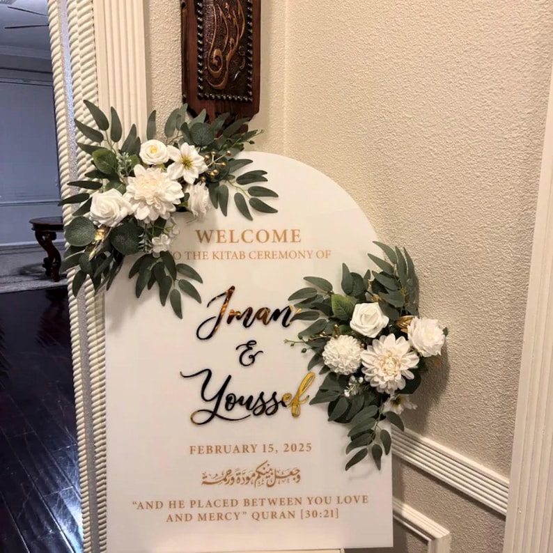 Puede incluir: Un cartel de bienvenida blanco con letras doradas para una ceremonia de boda. El cartel dice "Bienvenido a la ceremonia del Kitab de Iman & Youssef, 15 de febrero de 2025." El cartel tambi&eacute;n incluye caligraf&iacute;a &aacute;rabe y una cita del Cor&aacute;n: "Y &Eacute;l puso entre vosotros el amor y la misericordia." Cor&aacute;n [30:21].