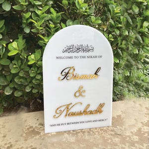 Może przedstawiać: Biały znak w kształcie łuku z złotymi literami. Znak głosi "Witamy na Nikah Bismah & Noushadh" i "I On umieścił między wami miłość i miłosierdzie".
