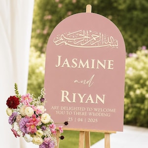 May include: An arched dusty rose wedding sign on a wooden easel featuring white Arabic calligraphy at the top, the names JASMINE and RIYAN in white serif font, and the text ARE DELIGHTED TO WELCOME YOU TO THERE WEDDING 25 | 04 | 2025.