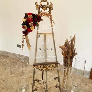 Puede incluir: Un caballete dorado con un letrero de acr&iacute;lico transparente que dice "Adam y Zara" en letras doradas. El letrero tambi&eacute;n incluye caligraf&iacute;a &aacute;rabe y la frase "Y te creamos en parejas". El caballete est&aacute; decorado con flores secas y un ramo de rosas rojas y doradas.