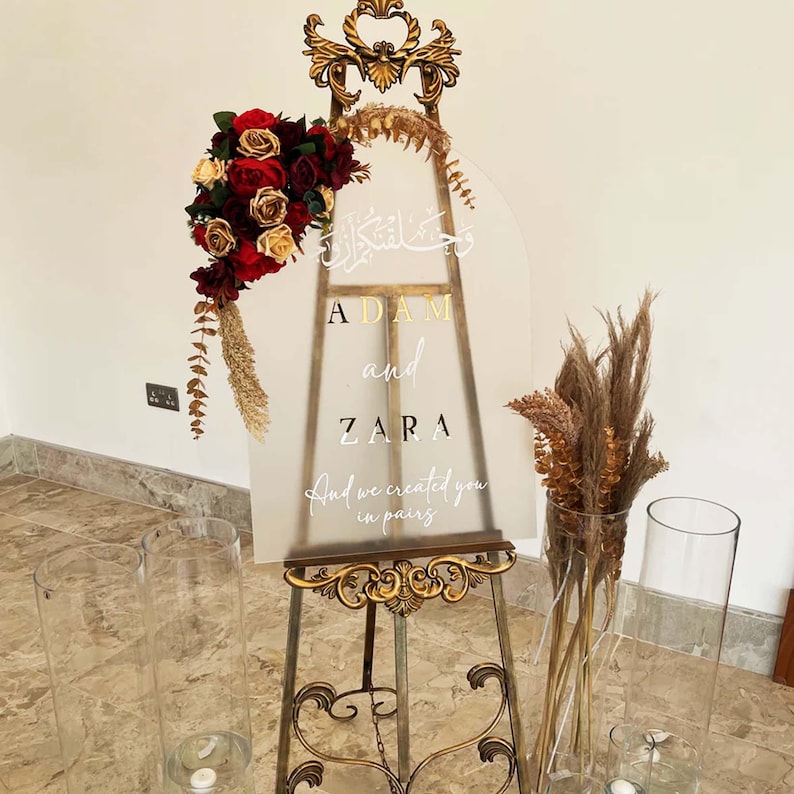 Puede incluir: Un caballete dorado con un letrero de acr&iacute;lico transparente que dice "Adam y Zara" en letras doradas. El letrero tambi&eacute;n incluye caligraf&iacute;a &aacute;rabe y la frase "Y te creamos en parejas". El caballete est&aacute; decorado con flores secas y un ramo de rosas rojas y doradas.