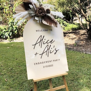 Puede incluir: Un cartel blanco en forma de arco con el texto "Welcome to Alice + Ali's Engagement Party 2-10-2022" en un caballete de madera. El cartel est&aacute; decorado con flores secas y hierba de la pampa.