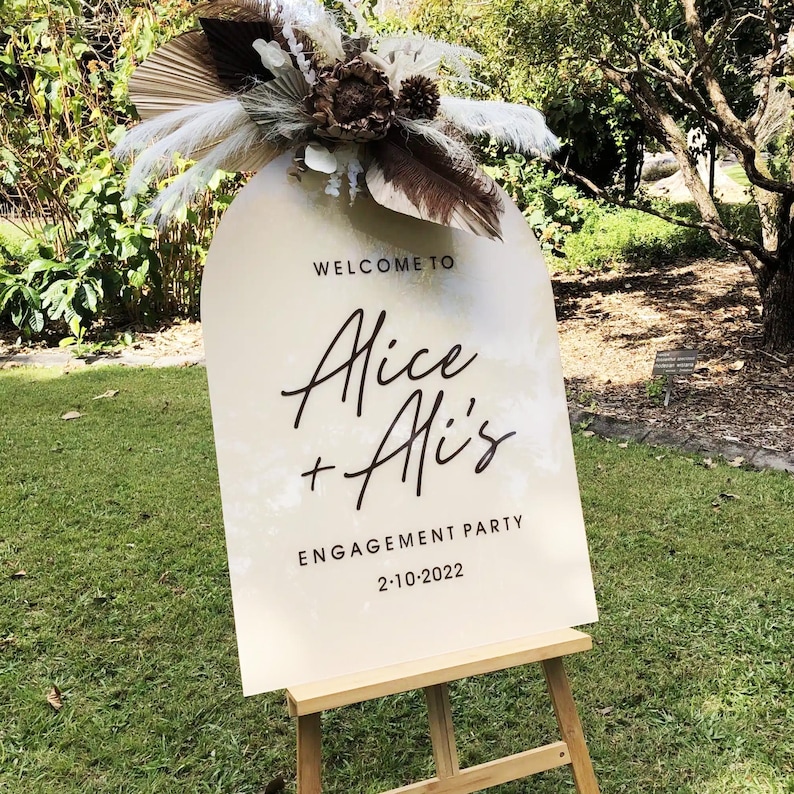 Puede incluir: Un cartel blanco en forma de arco con el texto "Welcome to Alice + Ali's Engagement Party 2-10-2022" en un caballete de madera. El cartel est&aacute; decorado con flores secas y hierba de la pampa.
