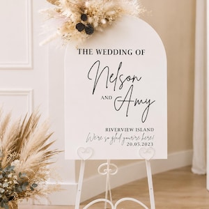 May include: An arched white wedding sign featuring black text that reads THE WEDDING OF Nelson AND Amy, RIVERVIEW ISLAND, We're so glad you're here! and 20.05.2023. The sign is decorated with dried beige grasses and sits on a white metal easel.