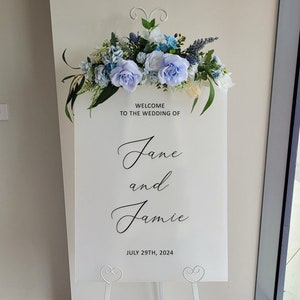 Puede incluir: Se&ntilde;al de bienvenida de boda blanca con flores azules y blancas en la parte superior. La se&ntilde;al dice "Bienvenido a la boda de Jane y Jamie 29 de julio de 2024".