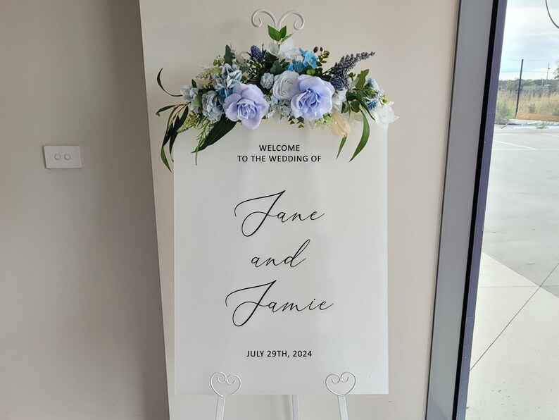 Puede incluir: Se&ntilde;al de bienvenida de boda blanca con flores azules y blancas en la parte superior. La se&ntilde;al dice "Bienvenido a la boda de Jane y Jamie 29 de julio de 2024".