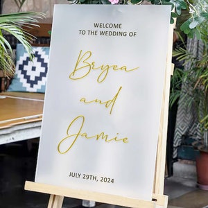 Puede incluir: Se&ntilde;al de bienvenida de boda blanca con letras doradas. La se&ntilde;al dice "Bienvenido a la boda de Bryea y Jamie 29 de julio de 2024".