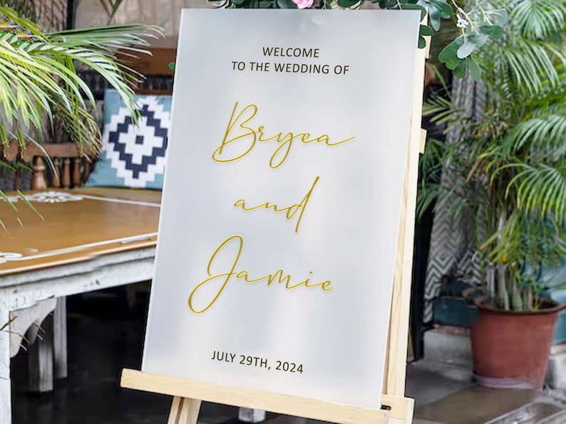 Puede incluir: Se&ntilde;al de bienvenida de boda blanca con letras doradas. La se&ntilde;al dice "Bienvenido a la boda de Bryea y Jamie 29 de julio de 2024".