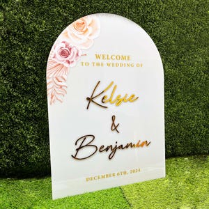 Puede incluir: Un cartel de bienvenida blanco en forma de arco con letras doradas. El cartel dice "Bienvenido a la boda de Kelse & Benjamin" y "Diciembre 6th, 2024". El cartel est&aacute; decorado con ilustraciones florales rosas y blancas.