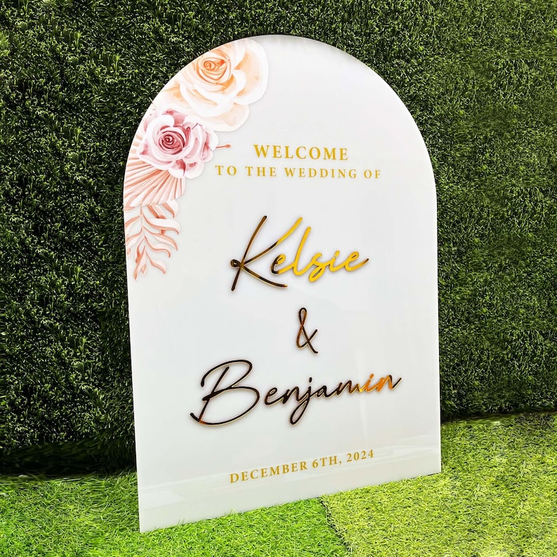 Puede incluir: Un cartel de bienvenida blanco en forma de arco con letras doradas. El cartel dice "Bienvenido a la boda de Kelse & Benjamin" y "Diciembre 6th, 2024". El cartel est&aacute; decorado con ilustraciones florales rosas y blancas.