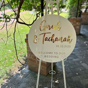 May include: White round wedding sign with gold lettering. The sign says "Girish & Tachanah" and "01.12.2023" with the words "Welcome to our Wedding" below. The sign is on a white easel.