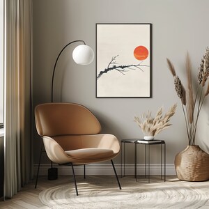 May include: A minimalist living room with a brown armchair, a floor lamp, a side table, and a framed print of a black branch with a red circle on a beige background. The room is decorated with dried pampas grass in a woven basket.