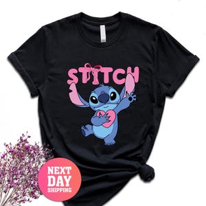 May include: Black t-shirt featuring the character Stitch from Lilo & Stitch. Stitch is blue and holding a pink heart. The word "STITCH" is printed in pink above Stitch's head.