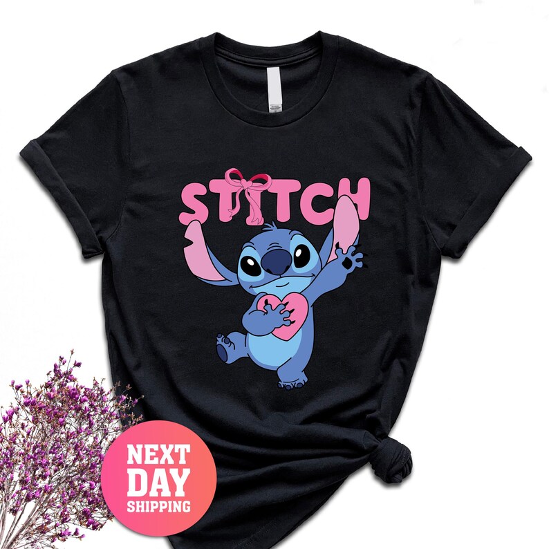 May include: Black t-shirt featuring the character Stitch from Lilo & Stitch. Stitch is blue and holding a pink heart. The word "STITCH" is printed in pink above Stitch's head.