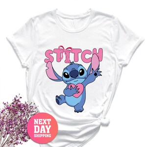 May include: White t-shirt with a cartoon image of Stitch holding a pink heart. The word "STITCH" is printed above the character in pink. A pink and red circular sticker with the words "NEXT DAY SHIPPING" is in the lower left corner.