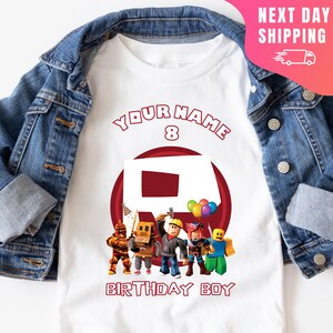 May include: White t-shirt with a red circle and the text "YOUR NAME 8" above it. Inside the circle is a red Roblox logo with a group of Roblox characters standing in front of it. The text "BIRTHDAY BOY" is below the circle.