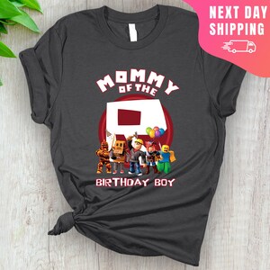 May include: A dark gray t-shirt with the text "Mommy of the" and a red Roblox "R" logo.  The shirt also features a group of Roblox characters standing in front of the "R".  The text "Birthday Boy" is below the characters.