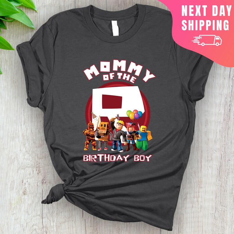 May include: A dark gray t-shirt with the text "Mommy of the" and a red Roblox "R" logo.  The shirt also features a group of Roblox characters standing in front of the "R".  The text "Birthday Boy" is below the characters.