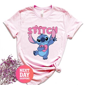 May include: Pale pink t-shirt with a cartoon character holding a heart. The word "STITCH" is printed above the character in pink. A round sticker with the text "NEXT DAY SHIPPING" is visible.