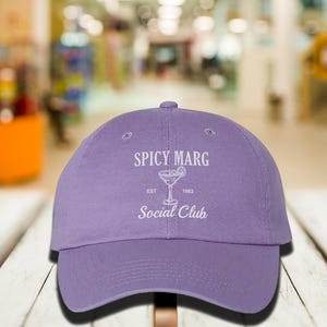 May include: A lavender baseball cap with the text "SPICY MARG Social Club" in white. The design includes a margarita glass graphic and the year "1983". The cap is made of a soft material and has a curved brim.